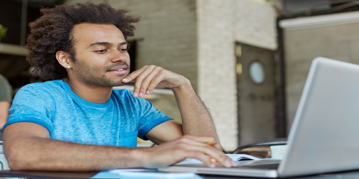 Pessoa com camiseta azul digitando no laptop enquanto faz anotações em um caderno sobre mesa de madeira
